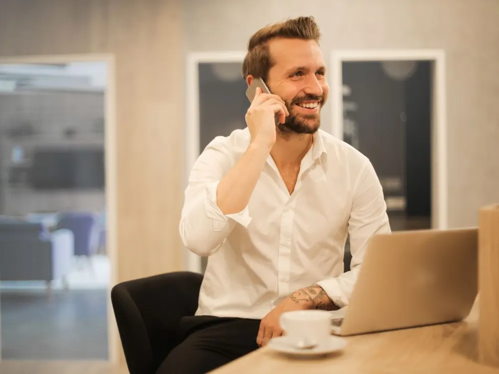 A smiling man in a white shirt talking on his phone at a desk with a laptop and a cup of coffee