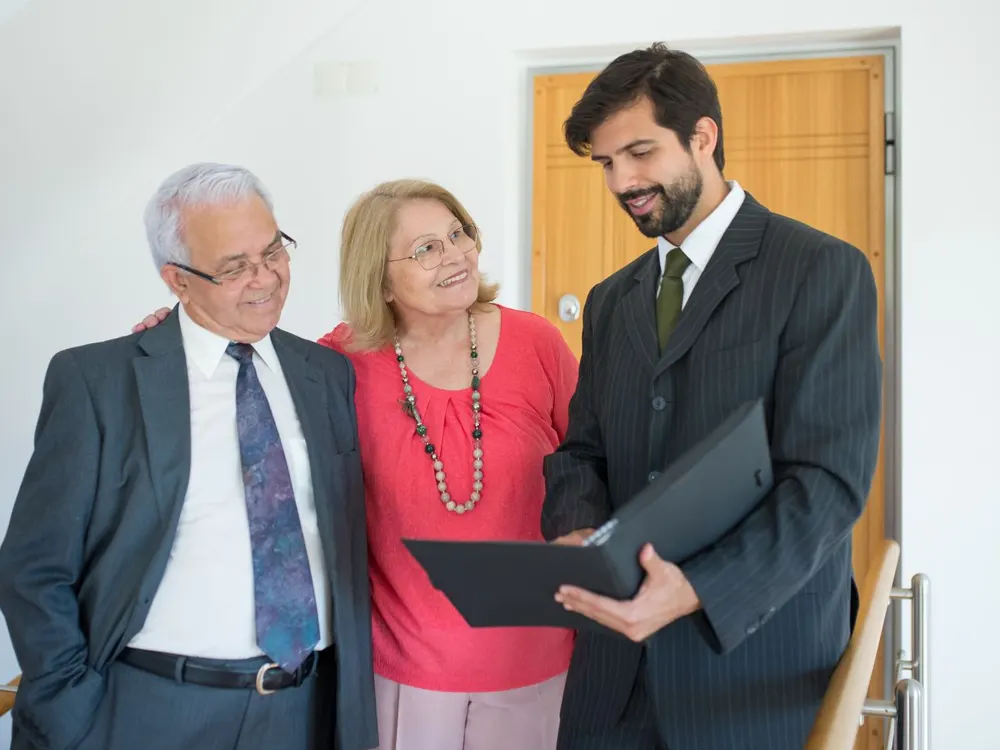A real estate agent in a suit holding a folder while talking with a smiling older couple at their front door