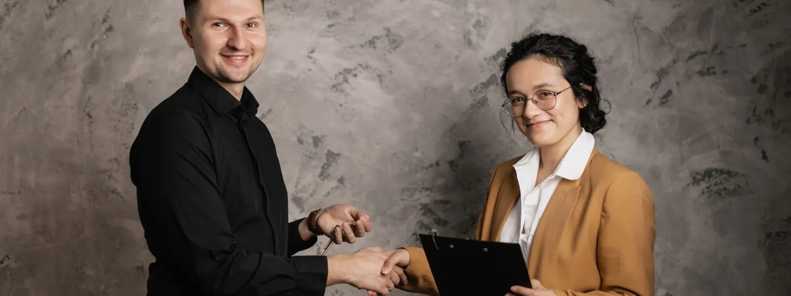 A smiling man and woman in business clothes shaking hands, the woman holding a clipboard
